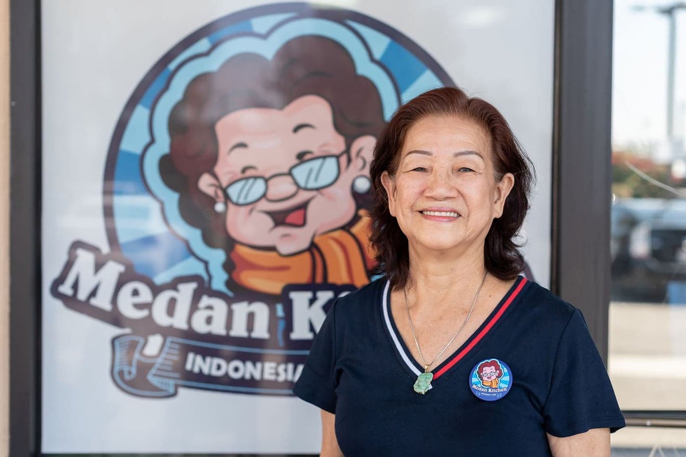 Siu Chen standing outside Medan Kitchen, beside the window decal of the shop’s logo