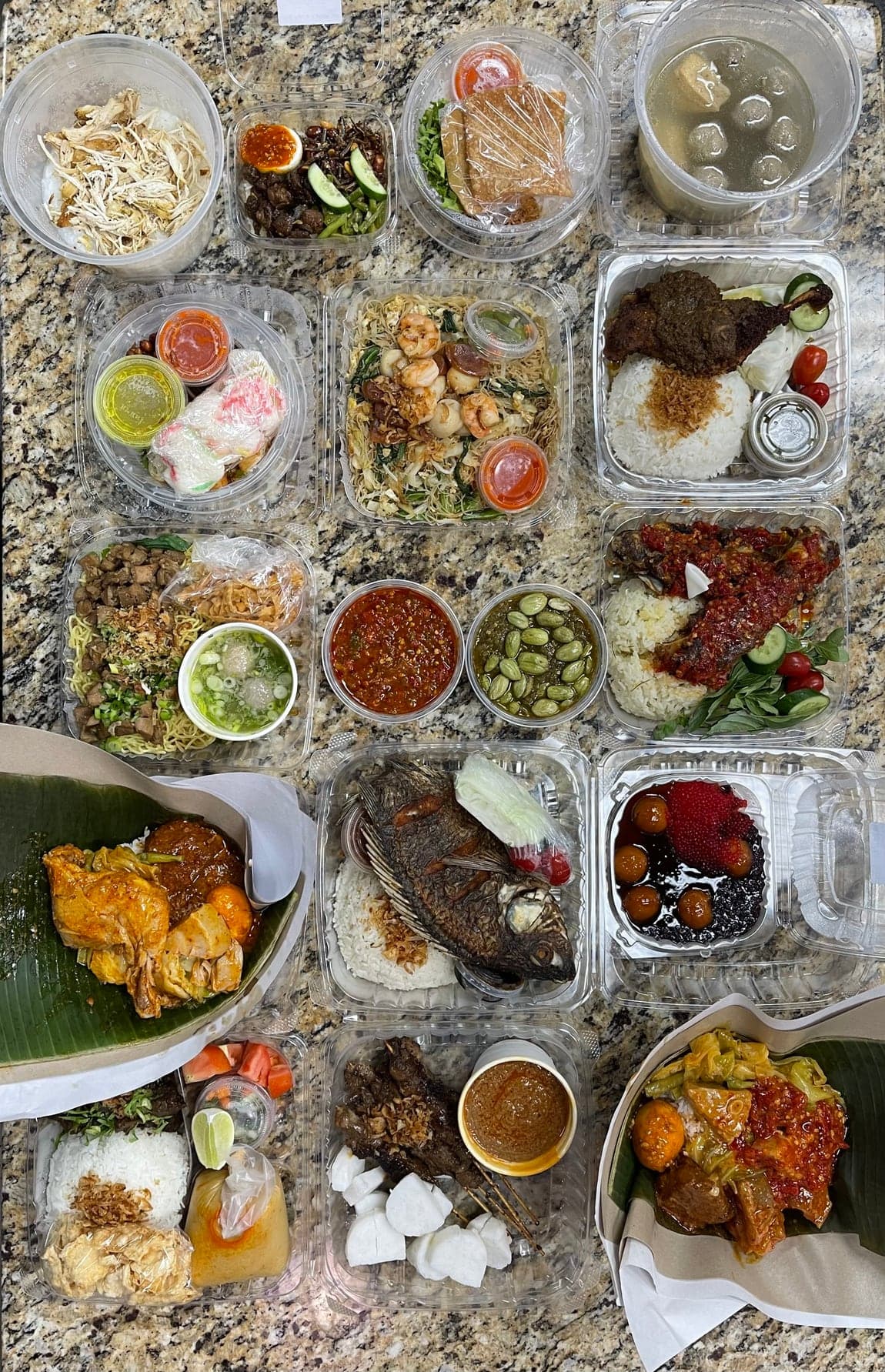 Around sixteen packed takeout containers laid out in a grid on the counter: Nasi Kapau and Nasi Padang, a whole fried fish, Bakso Campur in its broth, Bihun Goreng Seafood, Sate Ayam, two sambals and Bubur Madura